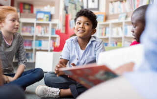 Multiethnic group of kids sitting on floor in circle around the teacher and listening a magazine reading